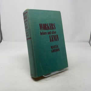 May include: A hardcover book titled "WORKERS before and after LENIN" by Manya Gordon. The book has a teal cover with red lettering. The spine also displays the title and author. The book is propped up by a white stand.