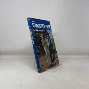 May include: A blue hardback book titled "The Gangster Film" by John Baxter. The cover features a colour image of a man and woman. The book is displayed on a clear stand. The title is in white text.