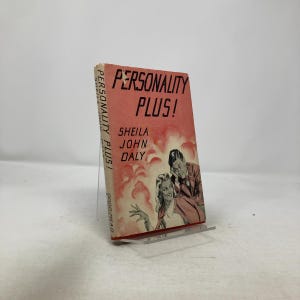 May include: A vintage book titled "Personality Plus!" by Sheila John Daly. The cover is a faded red with black text and an illustration of a man and woman. The book is displayed on a clear stand.