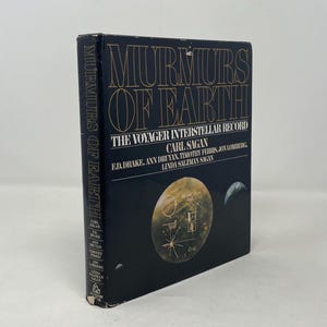 May include: A hardcover book titled "Murmurs of Earth: The Voyager Interstellar Record" by Carl Sagan. The cover is dark blue with gold lettering and a graphic of a planet and a crescent moon. The book's spine also has text.