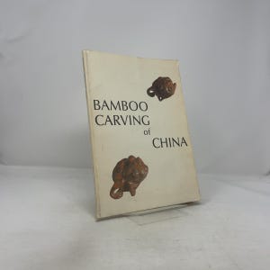 Könnte beinhalten: Ein Vintage-Buch mit dem Titel "Bamboo Carving of China" mit cremefarbenem Einband. Der Einband zeigt den Titel in schwarzer Schrift und zwei braune geschnitzte Figuren. Das Buch steht auf einem transparenten Ständer.