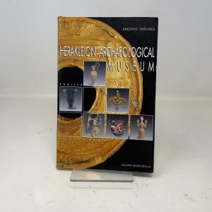 May include: A book titled "Herakleion Archaeological Museum" with the text "English" and "Visitor's Guide." The cover features a gold-colored design and images of artifacts. The book is standing on a clear plastic stand.