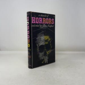 Puede incluir: Un libro de tapa dura vintage titulado "A Chamber of Horrors" con una cubierta negra y una ilustración de calavera. El título está en rosa y amarillo, con el nombre del autor, John Hadfield, debajo. El lomo del libro también muestra el título.