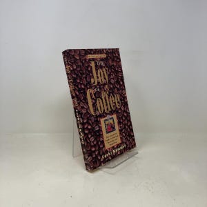 May include: A hardback book titled "The Joy of Coffee" is displayed on a clear stand. The cover features a background of coffee beans with the title in large, gold lettering. Additional text includes "Revised and Updated" and the author's name.