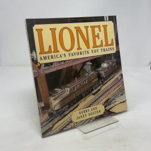 May include: A book titled "Lionel America's Favorite Toy Trains" featuring a brown and silver model train on tracks. The cover has the word "Lionel" in large orange letters. The book is propped up on a clear stand.