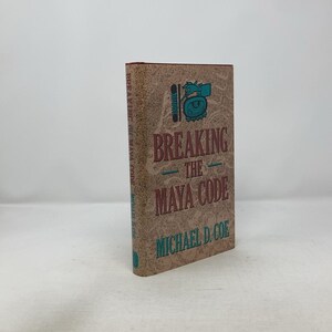 Puede incluir: Un libro de tapa dura titulado "Breaking the Maya Code" de Michael D. Coe. El libro tiene una cubierta de color beige con texto e ilustración turquesa y roja. El libro está de pie.