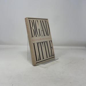 May include: A hardcover book titled "BIG AND LITTLE" by Botho Strauss. The book has a beige cover with black and red text. The book is propped up on a clear acrylic stand.