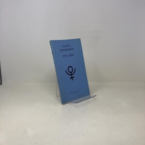 May include: A light blue book titled "PLUTO EPHEMERIS 1773-2000" with a black Pluto symbol. The book is displayed on a clear acrylic stand. The book's cover is slightly worn.