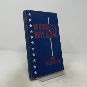 Puede incluir: Un libro de tapa dura azul titulado Wendell Willkie of Elwood con letras rojas. La portada presenta una columna vertical de estrellas blancas a lo largo del borde izquierdo y una fina línea vertical blanca que separa el texto. El libro está de pie.