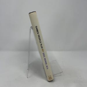 Könnte beinhalten: Ein Buch mit dem Titel "Games People Play" von Eric Berne, M.D. Das Buch hat einen hellbeigen Einband mit schwarzem Text. Es wird auf einem transparenten Kunststoffständer präsentiert, wobei der Buchrücken sichtbar ist.