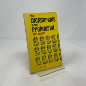 May include: A yellow book titled "The Dictatorship of the Proletariat" by Karl Kautsky. The book cover features the title in black and a series of portraits. The book is propped up on a clear stand.