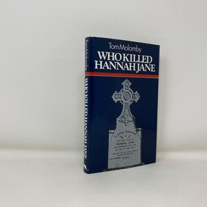 Puede incluir: Un libro azul con el título "Who Killed Hannah Jane" de Tom Malomby. La portada presenta una ilustración en blanco y negro de una lápida con una cruz.
