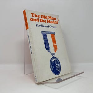 Può includere: Un libro bianco con un'illustrazione arancione e blu di una medaglia sulla copertina. Il titolo del libro è "Il vecchio e la medaglia" di Ferdinand Oyono.