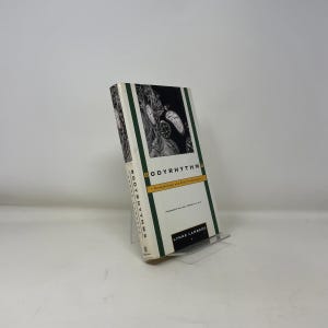 Könnte beinhalten: Ein Hardcover-Buch mit dem Titel "Bodyrhythms" steht auf einem transparenten Ständer. Das Buch hat einen weißen Einband mit einer Schwarz-Weiß-Illustration und grünen Rändern. Der Name des Autors, Lynne Lamberg, ist unten aufgedruckt.