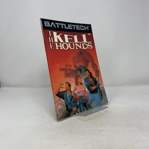 Op de afbeelding: Een paperback boek getiteld "BattleTech: The Kell Hounds" wordt op een transparante standaard getoond. De cover bevat illustraties van drie mensen en een grote robot tegen een rode achtergrond. De titel van het boek staat in witte en oranje letters.