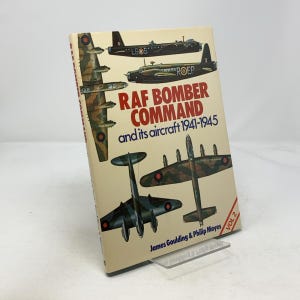 Puede incluir: Un libro antiguo titulado "RAF Bomber Command and its aircraft 1941-1945" con ilustraciones de aviones militares. La portada es beige con texto e imágenes en rojo, blanco y negro. El libro está apoyado en un soporte de plástico transparente.