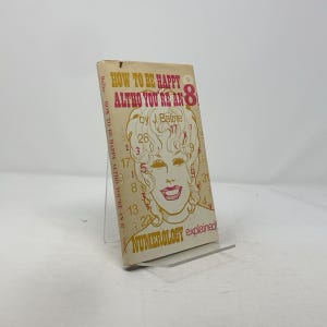 May include: A vintage paperback book titled "HOW TO BE HAPPY ALTHO YOU'RE AN 8" by J. Baline. The cover features a woman's face with numbers and the words "NUMEROLOGY explained!" in a retro design. The book is displayed on a clear stand.