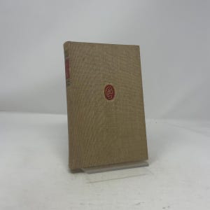 May include: A hardcover book with a tan linen cover. The spine has red text, and the front cover features a red circular emblem. The book is displayed on a clear acrylic stand.