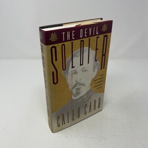 Op de afbeelding: Een hardcover boek getiteld The Devil Soldier van Caleb Carr. De kaft toont een sepia-kleurig portret van een man met een snor, tegen een getextureerde beige achtergrond met een donkerrode balk bovenaan met de titel en Chinese tekens.