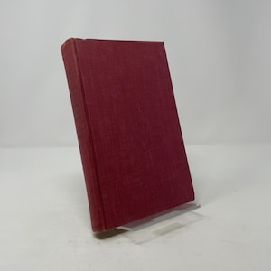 Könnte beinhalten: Ein Hardcover-Buch mit burgunderfarbenem Einband. Das Buch ist leicht angewinkelt, wodurch der Buchrücken und der strukturierte Stoff des Einbands sichtbar werden. Das Buch wird auf einem transparenten Acrylständer präsentiert.