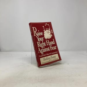 May include: A hardcover book titled "Raise Your Right Hand Against Fear" by Sheldon Kopp. The book has a red cover with white text and a hand illustration. The book is displayed on a clear stand.