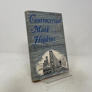 May include: A hardcover book titled Controversial Mark Hopkins, the Great Swindle of American History by Estelle Latta, Revised Edition. The grey dust jacket features blue text and a black and white illustration of a steam train and a bridge.
