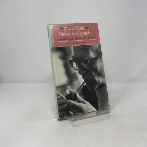 May include: A paperback book titled "Ordeal by Labyrinth" by Mircea Eliade. The cover features a black and white image of a man smoking a pipe. The book is propped up on a clear plastic stand.