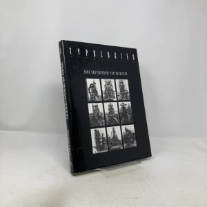 May include: A black book titled "Typologies" with the subtitle "Nine Contemporary Photographers." The cover features a grid of nine black and white photographs. The book is displayed on a clear stand.