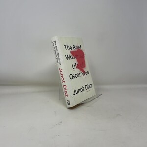 Puede incluir: Un libro titulado "La breve y maravillosa vida de Oscar Wao" de Junot Díaz. El libro tiene una cubierta blanca con texto rojo y negro y un gráfico rojo. El libro se muestra en un soporte transparente.