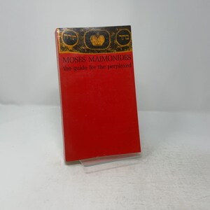 May include: A red book titled Moses Maimonides The Guide for the Perplexed. The top section features a black band with gold Hebrew text and a central gold crown icon. The cover is solid red with black text detailing the author and title.