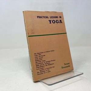 May include: A vintage book titled "Practical Lessons in Yoga" with a tan cover and green horizontal stripe. The book's title and author, Swami Sivananda, are printed in dark blue. The book is propped up on a clear acrylic stand.