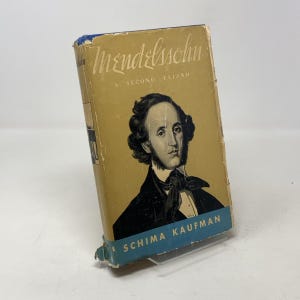 Puede incluir: Un libro antiguo titulado "Mendelssohn A Second Elijah" con un retrato de un hombre en la portada. El libro tiene una cubierta de color beige con letras azules y doradas. El libro está ligeramente desgastado, mostrando signos de edad.