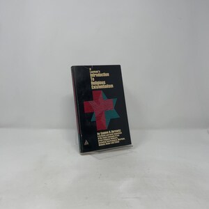 Puede incluir: Un libro negro titulado "A Layman's Introduction To Religious Existentialism" con un diseño de cruz y estrella rojo y verde azulado. El libro está apoyado en un soporte transparente sobre un fondo blanco.