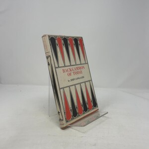 May include: A vintage book titled "Backgammon of Today" by John Longacre. The book cover features a backgammon board design with red and black triangles. The book is displayed on a clear acrylic stand against a white background.