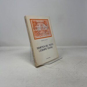 Könnte beinhalten: Ein Buch mit dem Titel "Semiotica del testo L'esempio teatro" von Franco Ruffini. Das Buch hat einen weißen Einband mit orangefarbenem und schwarzem Text und einer Illustration. Das Buch steht auf einem transparenten Ständer.