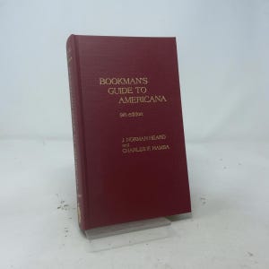 May include: A hardcover book titled Bookman's Guide to Americana, 9th edition, by J. Norman Heard and Charles F. Hamsa. The book features a solid deep red cover with gold lettering printed on the front and the spine, standing upright on a base.