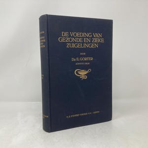 Può includere: Un libro rilegato in blu intitolato "De Voeding Van Gezonde En Zieke Zuigelingen" di Dr. E. Gorter, 8a edizione. La copertina del libro presenta un'illustrazione dorata di un serpente avvolto attorno a una lampada.