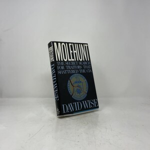 Könnte beinhalten: Ein gebundenes Buch mit dem Titel Molehunt: The Secret Search for Traitors That Shattered the CIA von David Wise. Der schwarze Schutzumschlag zeigt weißen Text und ein zentrales blau-goldenes Emblem der CIA mit einem roten Riss.