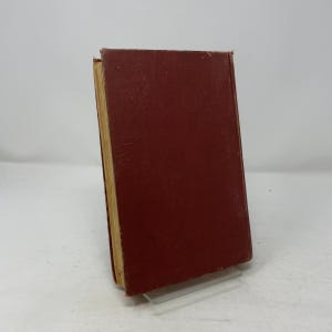 May include: A vintage hardcover book with a deep red cover. The book's pages have a yellowed appearance, suggesting age. The spine and edges show wear, indicating it has been well-read. The book is displayed on a clear acrylic stand.