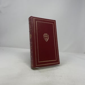 May include: A hardcover book with a deep red cover and gold-colored accents. The book features a gold-framed border and a gold emblem on the front cover. The spine has gold lettering. The book is standing upright.
