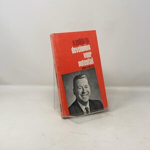 Può includere: Un libro rosso intitolato "a guide to developing your potential" di Robert L. Mox, Ph.D. La copertina del libro presenta una foto di un uomo in abito e cravatta che sorride.