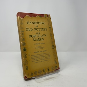 May include: A vintage book titled "Handbook of Old Pottery and Porcelain Marks" by C. Jordan Thorn. The cover is a faded yellow with black text and decorative elements. The book's spine is red, and the edges show signs of wear.