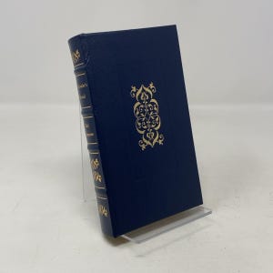Puede incluir: Un libro de tapa dura azul oscuro con letras doradas y elementos decorativos. El lomo y la portada presentan diseños dorados. El libro se exhibe sobre un soporte de plástico transparente, destacando su diseño y título.