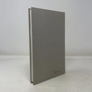 以下が含まれることがあります： グレーの表紙のハードカバーの本。背表紙が見え、白いページが表示されています。裏表紙には赤い文字が印刷されています。本は白い背景に立っています。