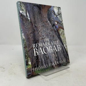 Op de afbeelding: Een hardcover boek getiteld The Remarkable Baobab door Thomas Pakenham, met op de kaft een grote, getextureerde, grijsbruine baobab-boomstam. Een persoon is zichtbaar terwijl deze in de boom klimt. Het boek staat op een doorzichtige acrylstandaard.