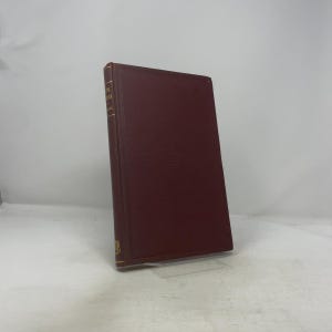 Puede incluir: Un libro de tapa dura burdeos con letras doradas en el lomo. El libro está de pie sobre un soporte transparente. El título y el nombre del autor son visibles. Los bordes de las páginas no son visibles.