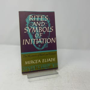 May include: A paperback book titled Rites and Symbols of Initiation by Mircea Eliade. The cover features a purple background with a teal stylized face illustration, a green lower section with white text, and a decorative border at the bottom.