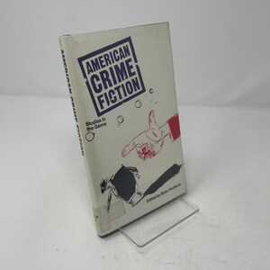 May include: A white paperback book titled American Crime Fiction: Studies in the Genre, edited by Brian Docherty. The cover features a blue rectangular border, a red and white line drawing of a hand pointing a gun, and a black silhouette figure.
