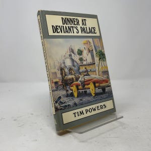 May include: A hardcover book titled Dinner at Deviant's Palace by Tim Powers. The cover features a color illustration of a horse-drawn carriage on a city street with buildings in the background. The title and author name are printed in black text.