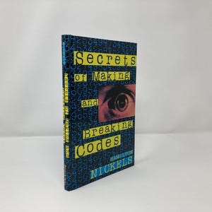 Puede incluir: Un libro de tapa dura titulado "Secrets of Making and Breaking Codes" de Hamilton Nickels. La portada es azul oscuro con texto amarillo y un primer plano de un ojo. Hay números impresos en la portada.